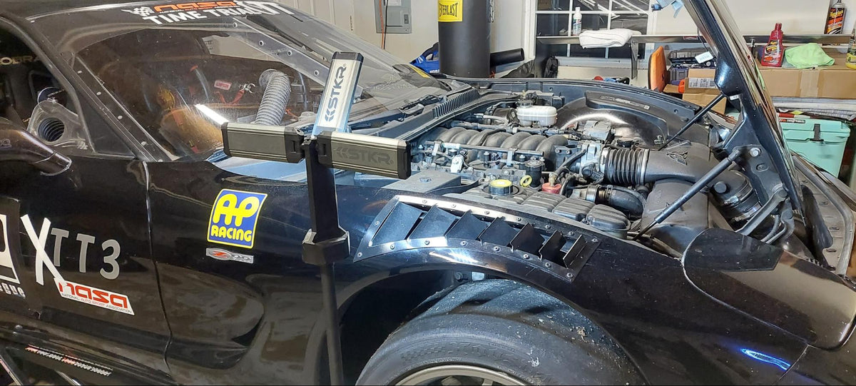 A TRi-MOBILE work light mounted on a tripod illuminates the open engine bay of a black race car in a garage, providing focused lighting for vehicle maintenance.
